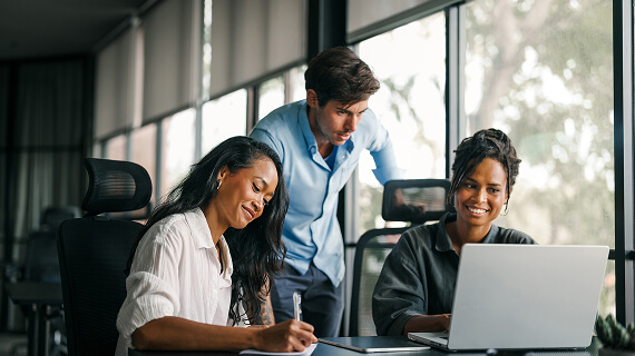 Man standing behind two woman who are taking notes and looking at a laptop screen
