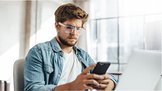 Casually dressed male with glasses sitting at his desk holding a mobile device