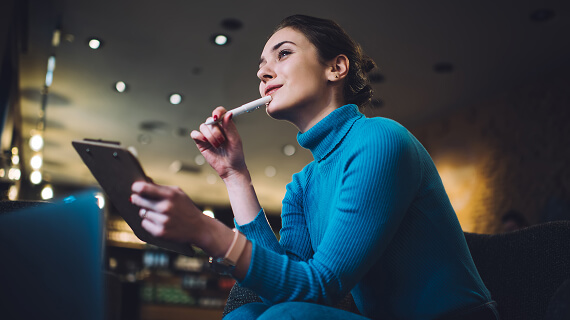 Seated woman holding a pen to her chin with a clipboard in the other hand